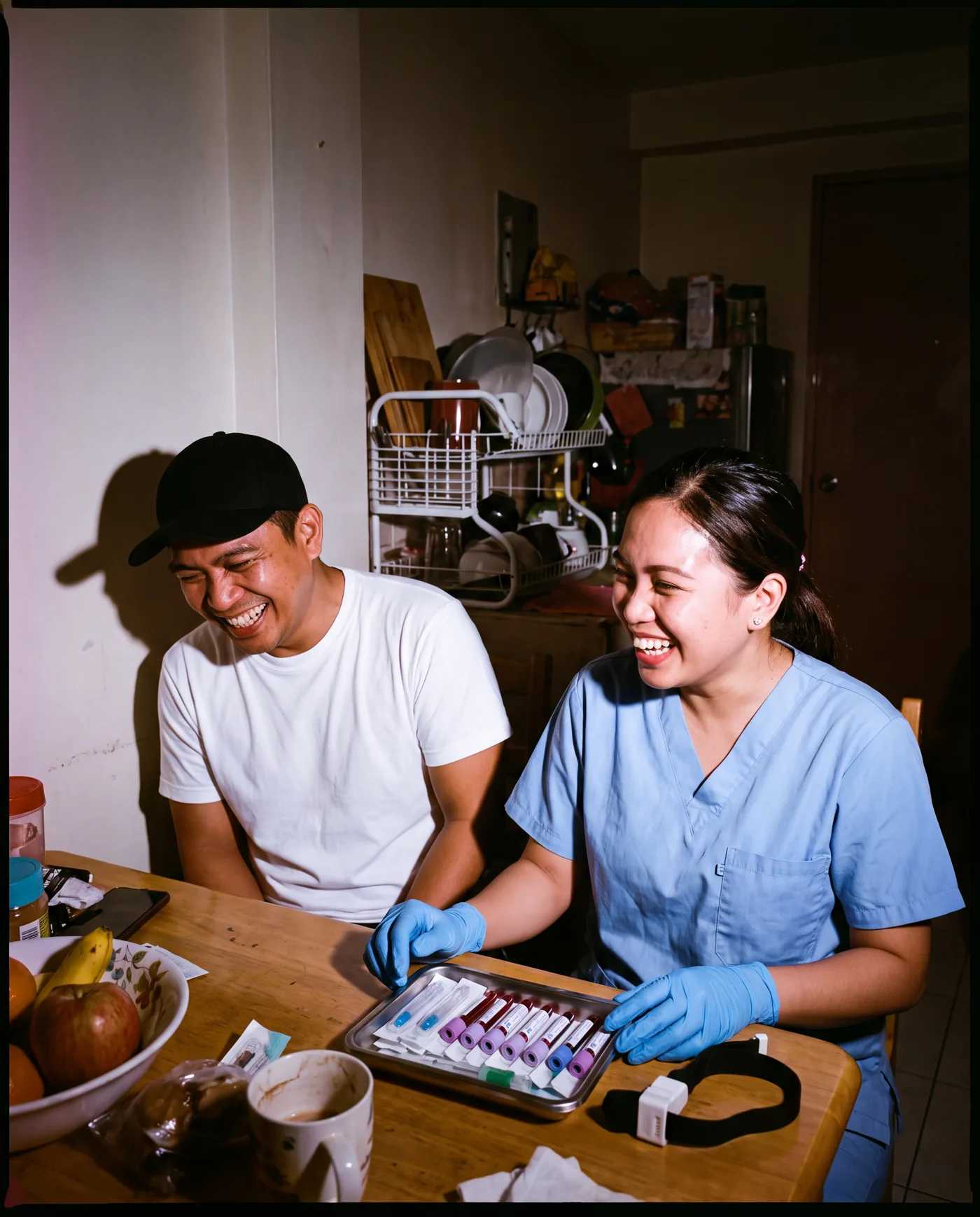 A licensed nurse and a patient laughing at his dining table in a Manila home, the sealed draw kit on the table between them.